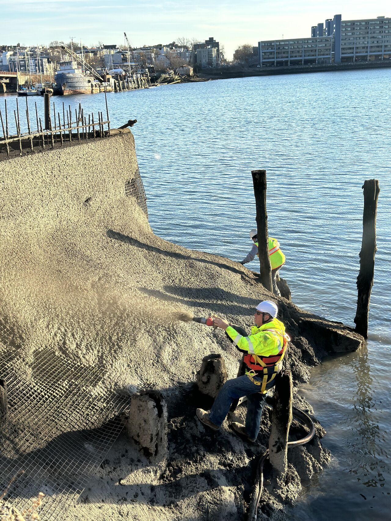 Two people in safety vests clean debris from a dock edge in a body of water, with buildings and boats in the background.