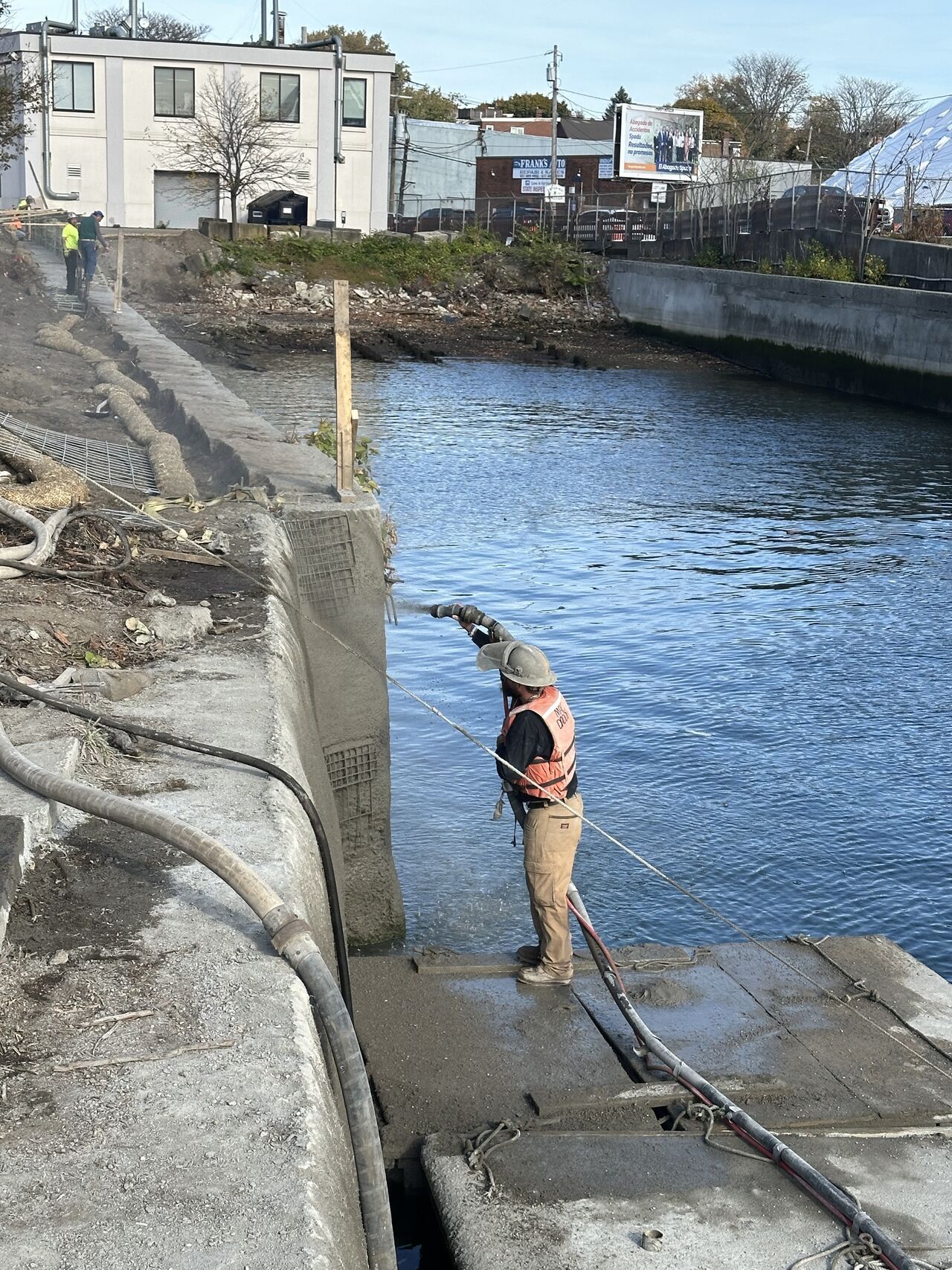 Man spraying water on a concrete wall near a waterway. A second worker stands in the background.