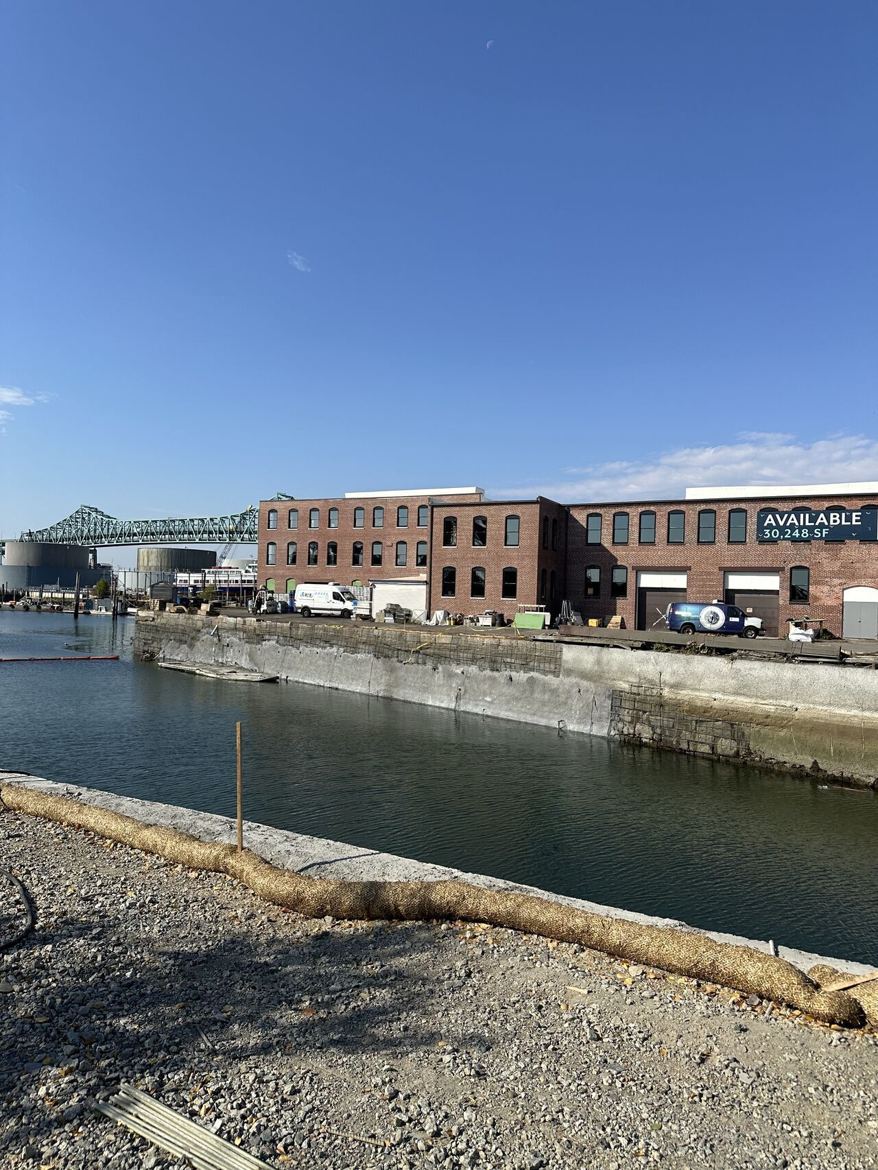 Brick building along a waterway with a bridge in the background under a blue sky.