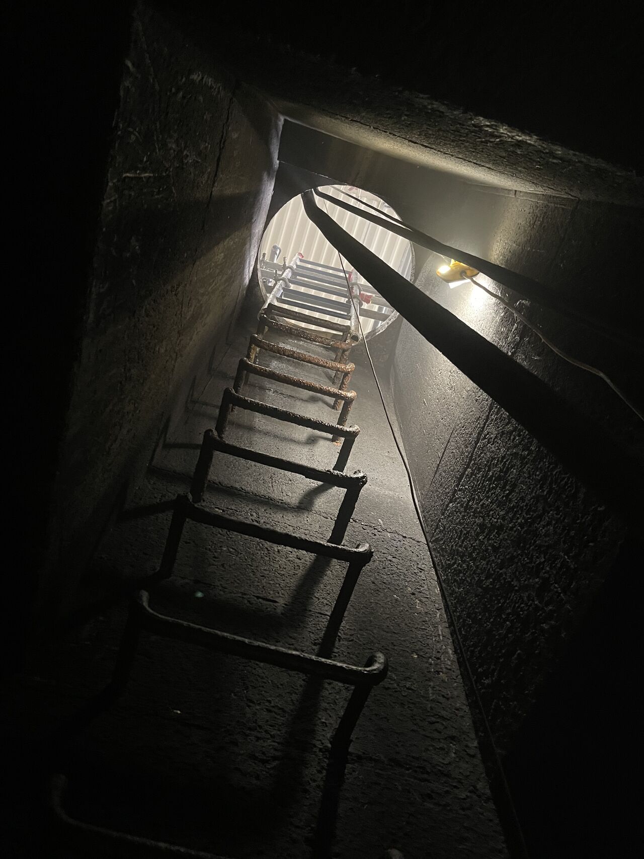 Ladder ascending into a dark shaft; circular opening at the top; a single light source illuminates the scene.