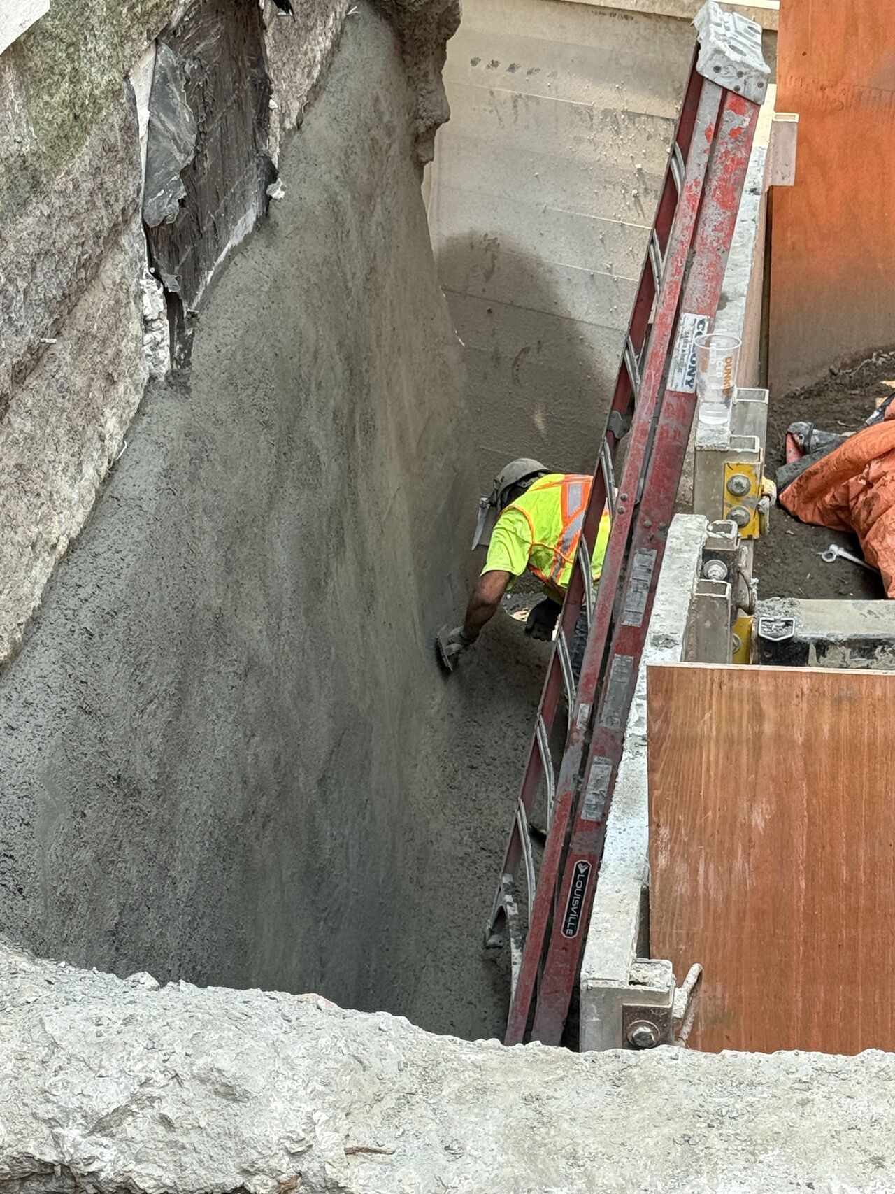 Construction worker applying concrete to a wall. A ladder and wood paneling are also in the shot.