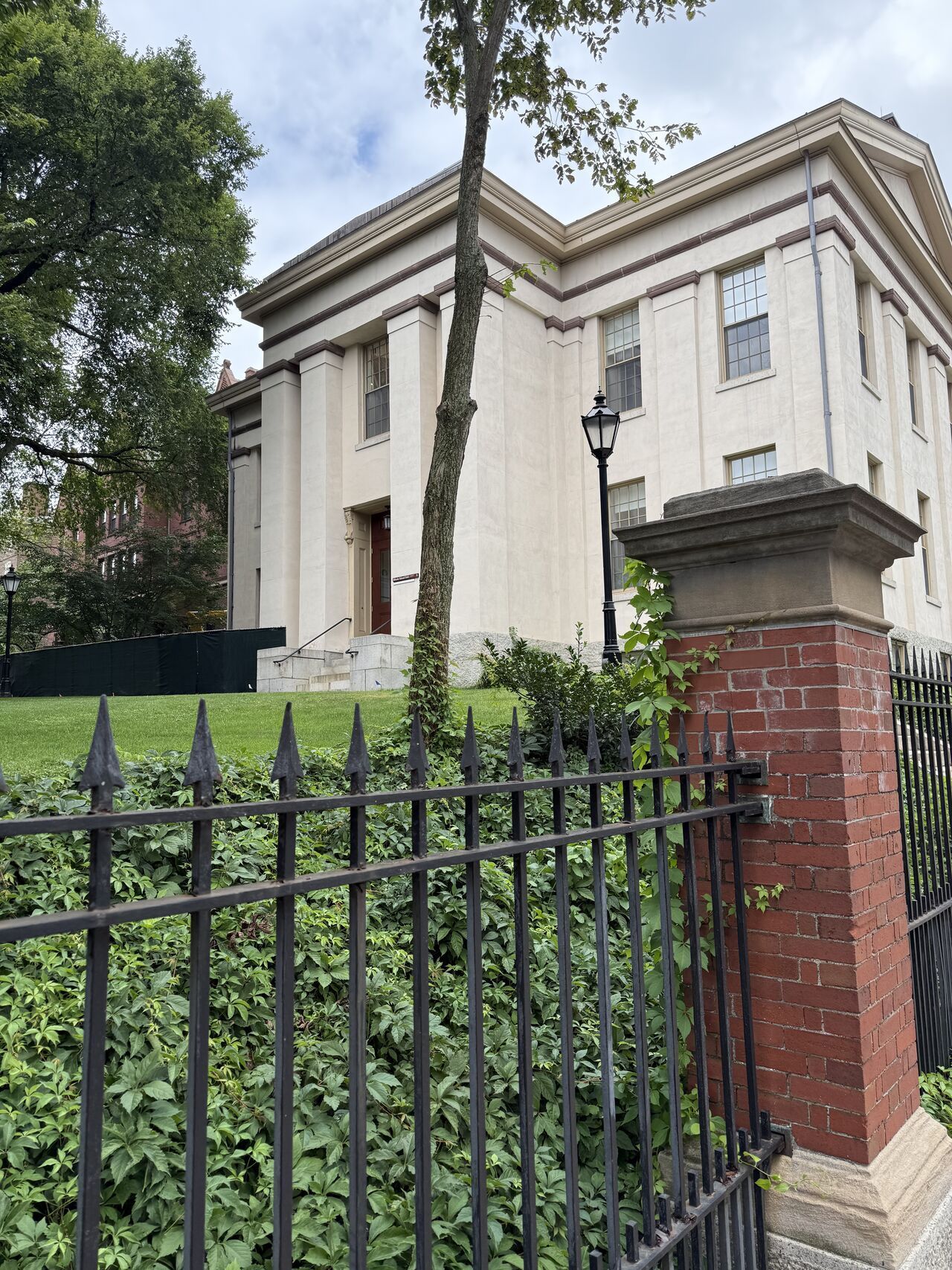 A building with columns behind a black fence, with a grassy hill and tree in front.