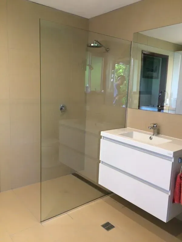Frameless glass shower screen in a beige tiled bathroom with a wall-mounted white vanity and mirror.