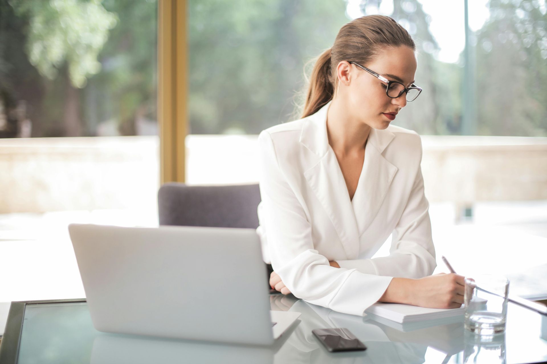 Woman in white blazer, glasses, and ponytail working at a desk with a laptop and writing notes.