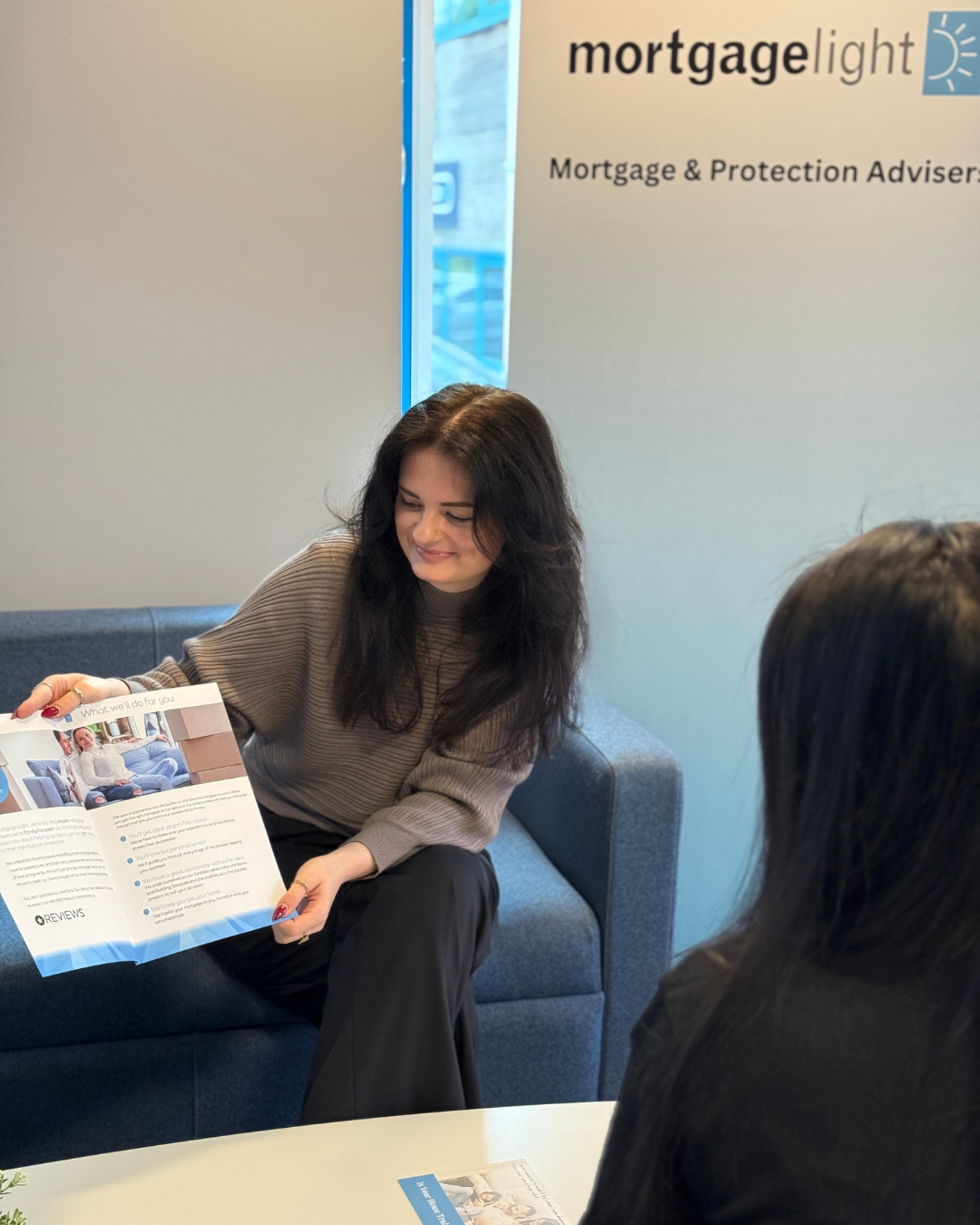 Woman with blonde hair smiles while talking to another woman, both seated on a blue couch.