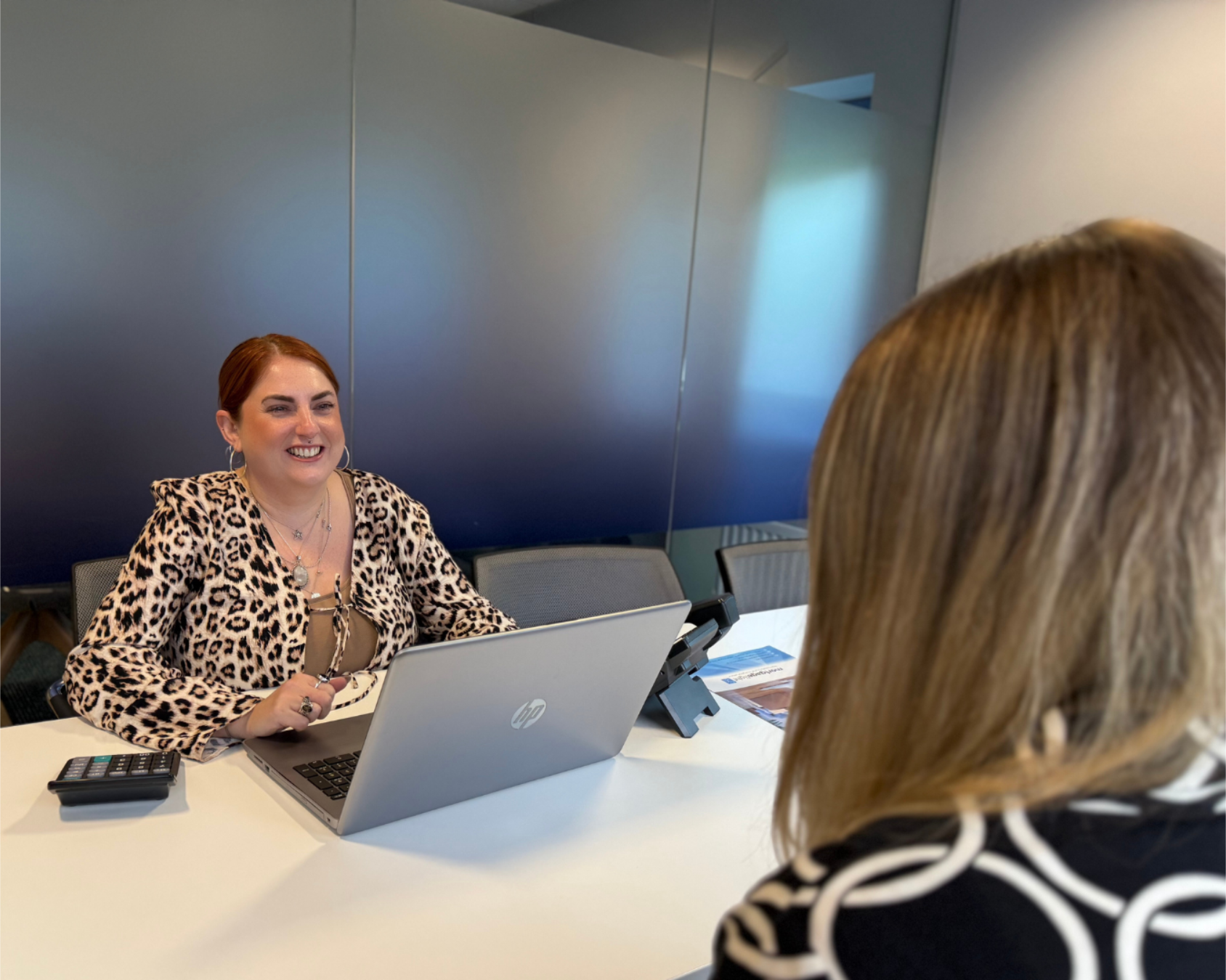 Woman with red hair smiles at a woman, seated at a table with a laptop. Inside office setting.