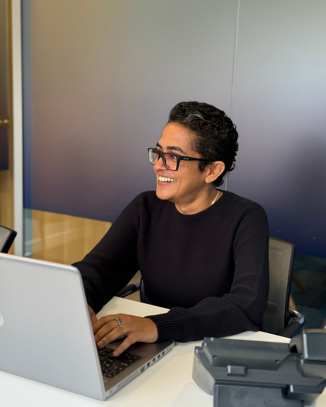 Woman with glasses smiles while typing on a laptop in an office.
