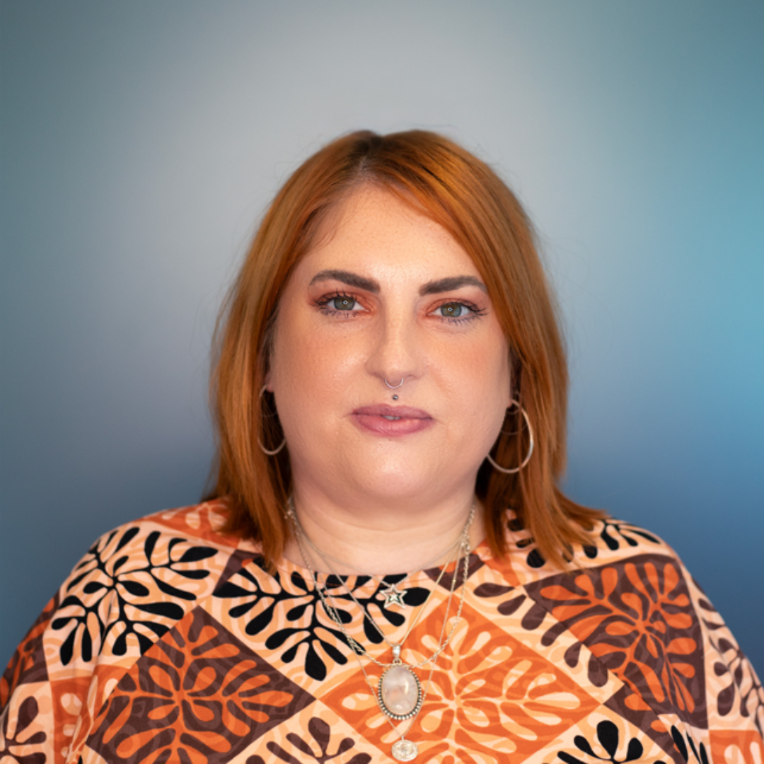 Woman with red hair and nose piercing, wearing a patterned top and jewelry, smiles against a blue gradient.