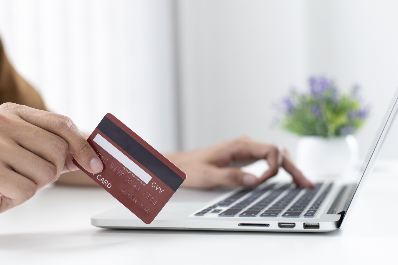 Person holding a red credit card, typing on a laptop at a white desk; flower pot in background.