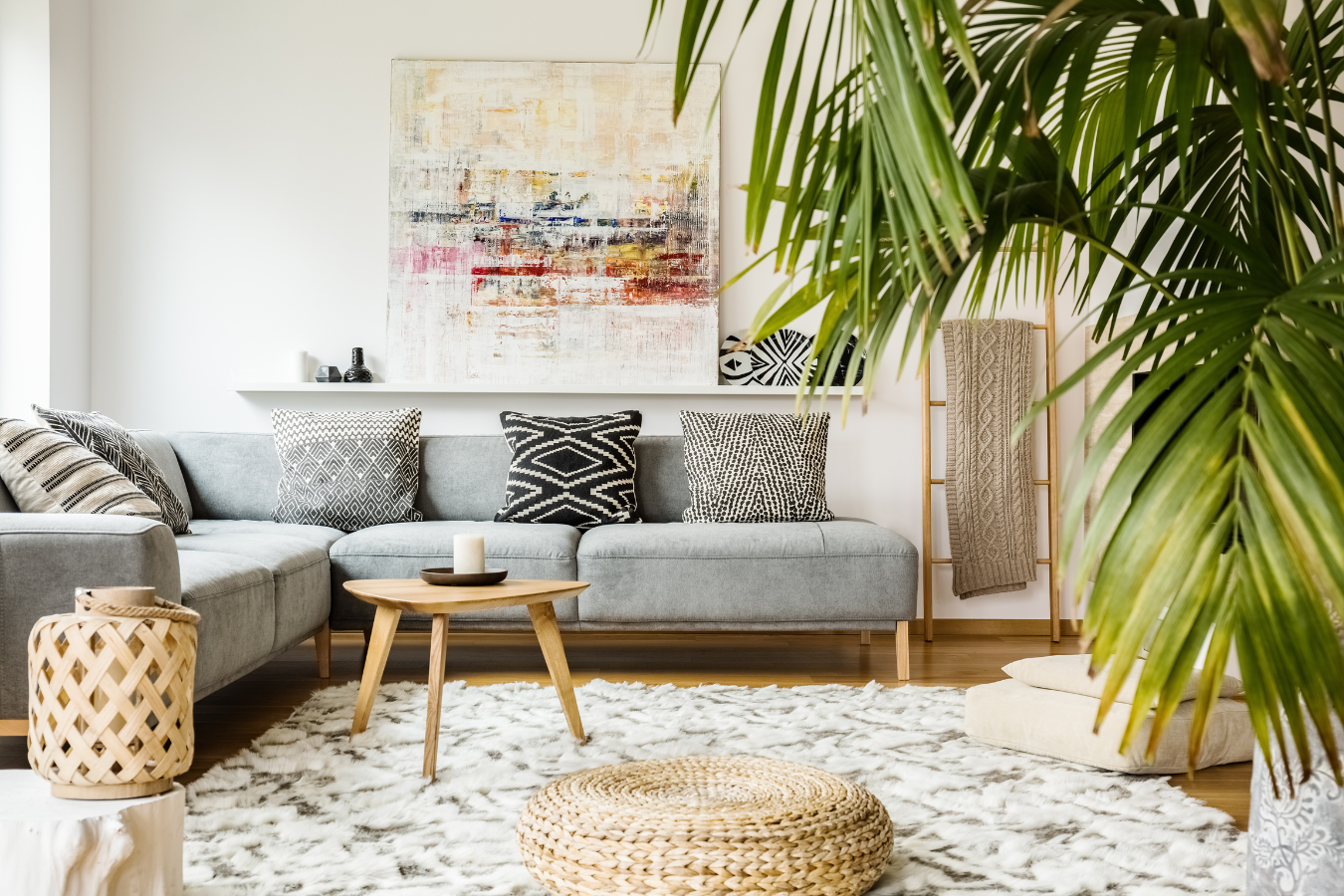 Cozy living room with grey sectional, patterned pillows, abstract art, rug, and large palm plant.
