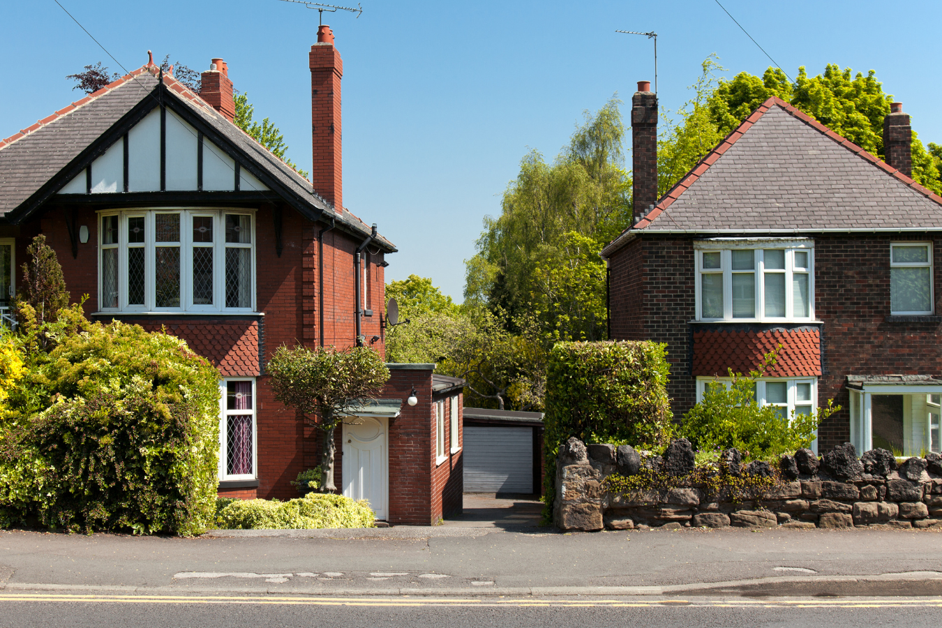 Brick houses with bay windows, chimneys, and a stone wall on a sunny street.