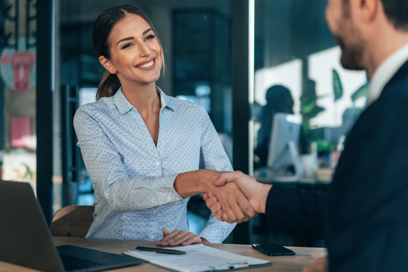 Woman in blue shirt shaking hands with a man in a suit, smiling, in an office.