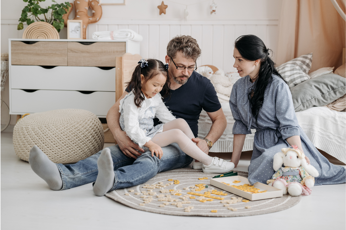 Family playing on the floor: father holding daughter, mother beside them, toys scattered on a rug in a light-filled room.