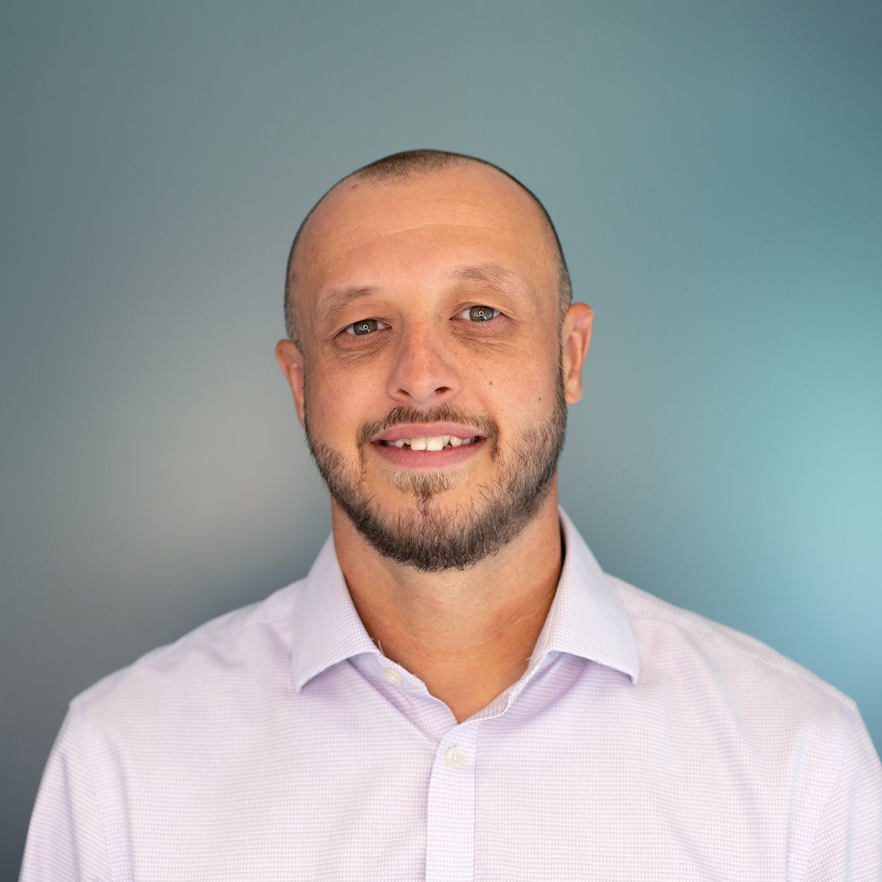 Man with short hair and beard smiling, wearing a light purple shirt, against a blue background.
