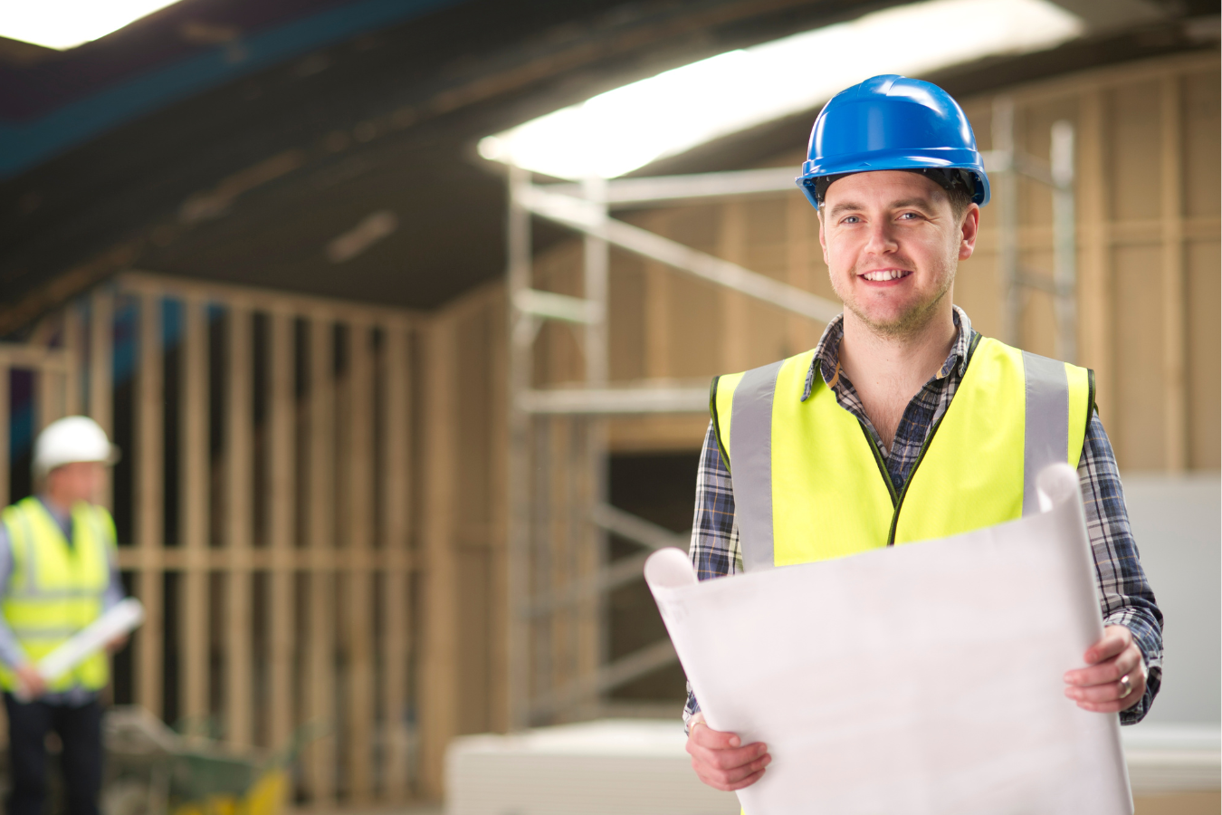 Construction worker smiling, holding blueprints, wearing hard hat and safety vest, inside building under construction.