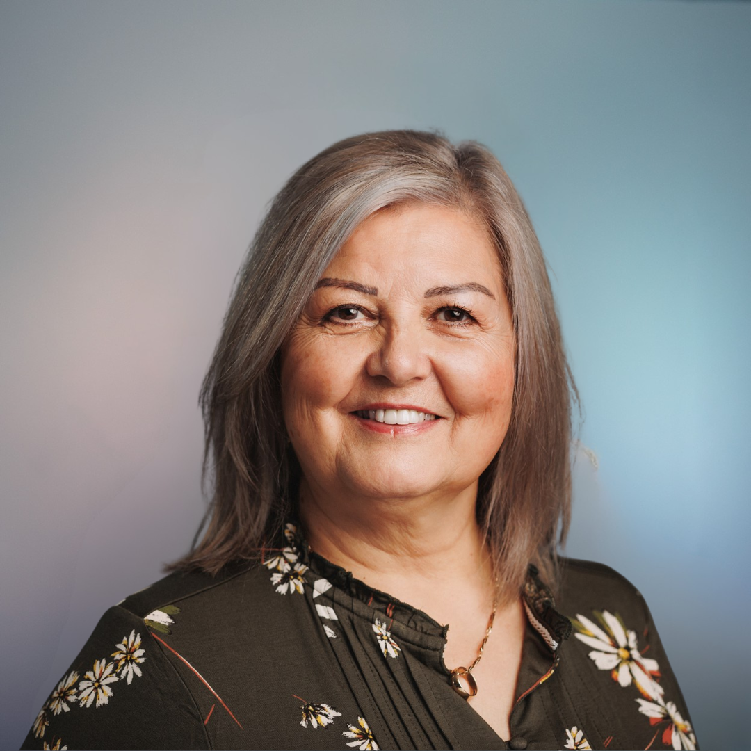 Woman with gray hair smiles at the camera, wearing a floral print top against a blurred blue background.