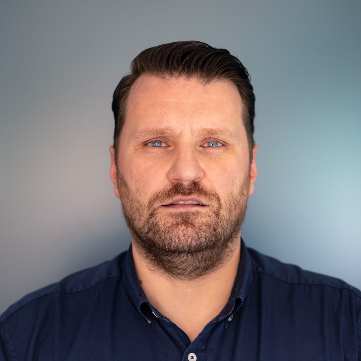 Man with brown hair and beard in a blue shirt, looking directly at the camera against a gradient blue background.