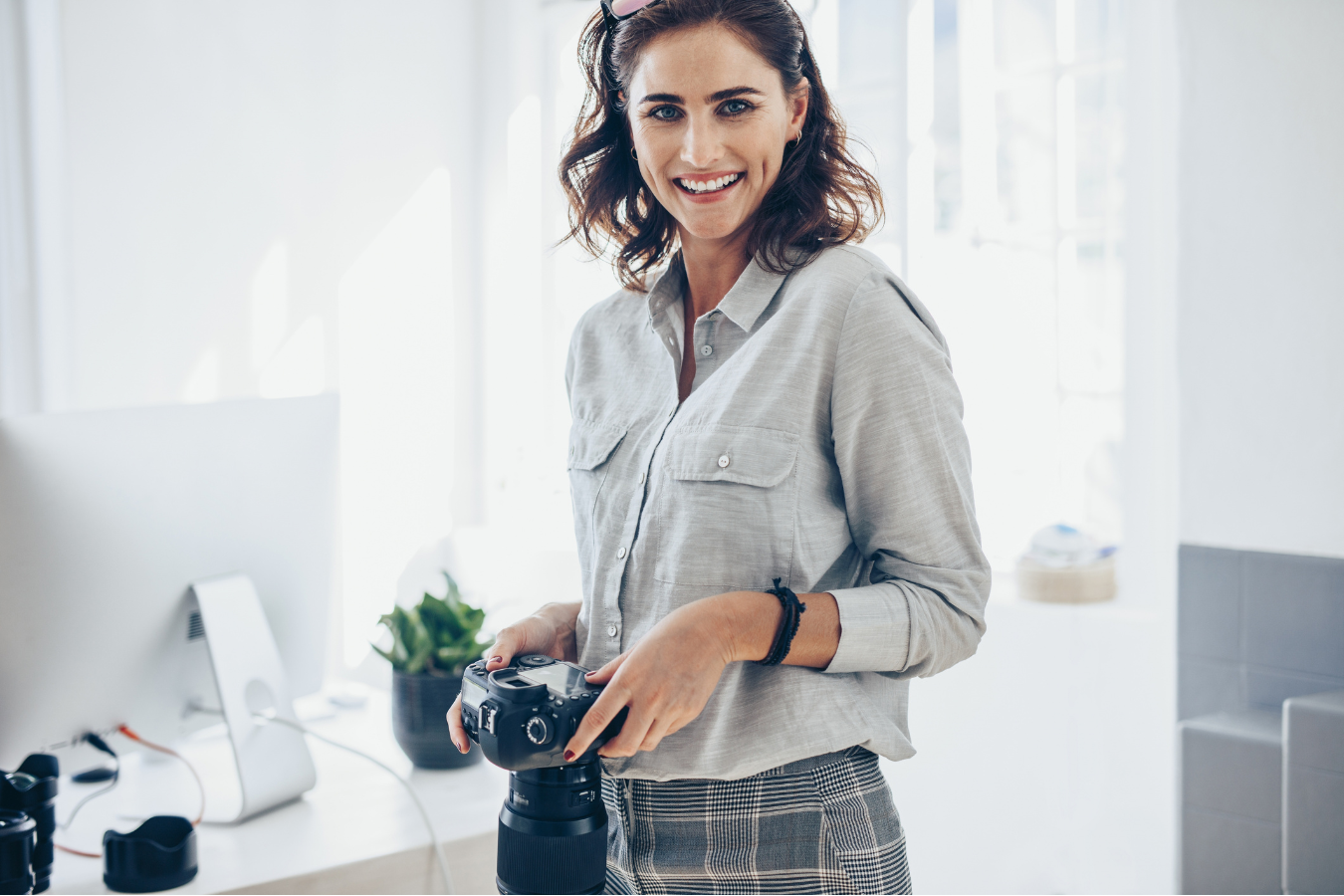 Woman with camera, smiling, in a bright room. Wearing a light grey shirt and holding a camera with a black lens.