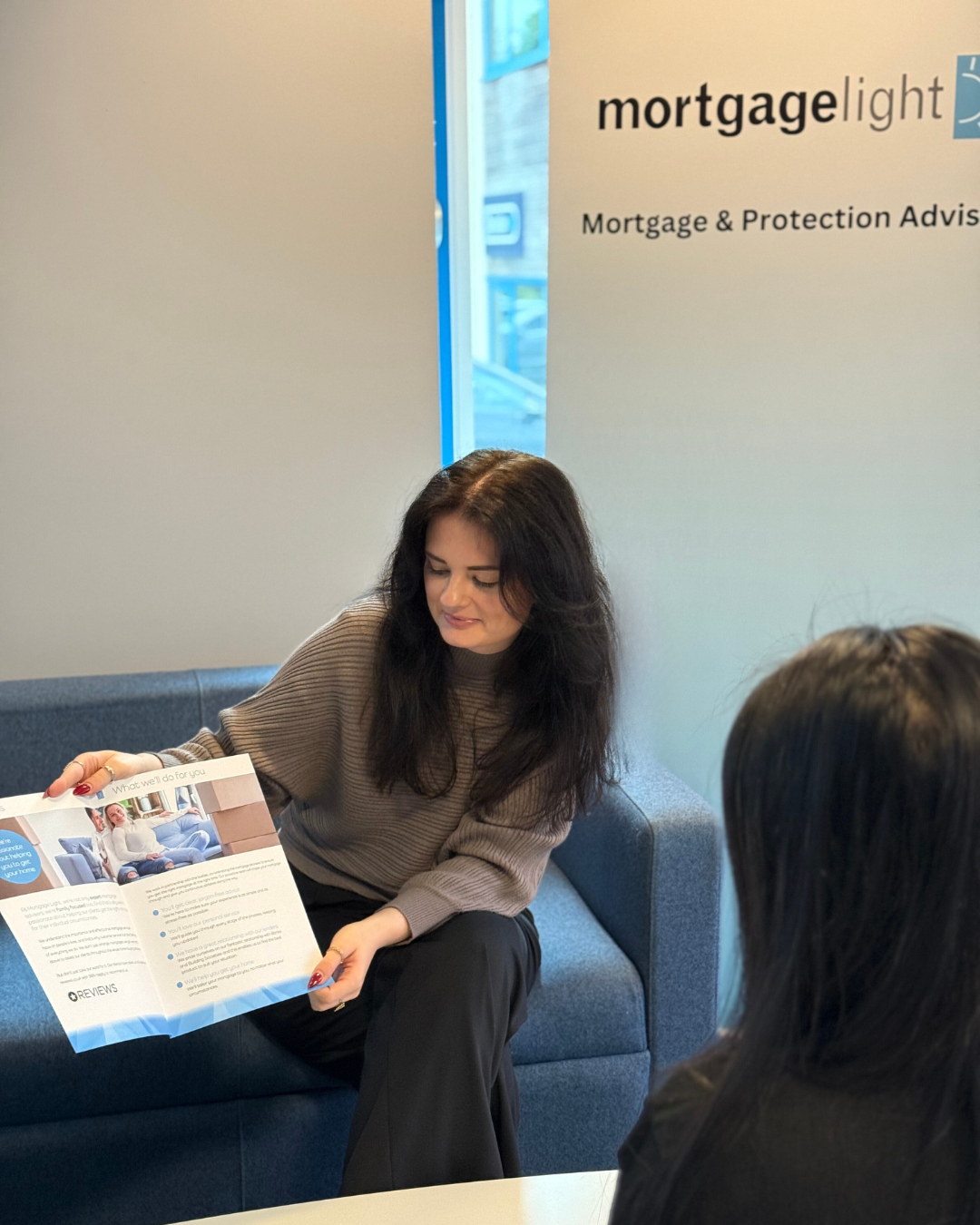 Woman in gray sweater shows a brochure to another person, in a mortgage office setting.