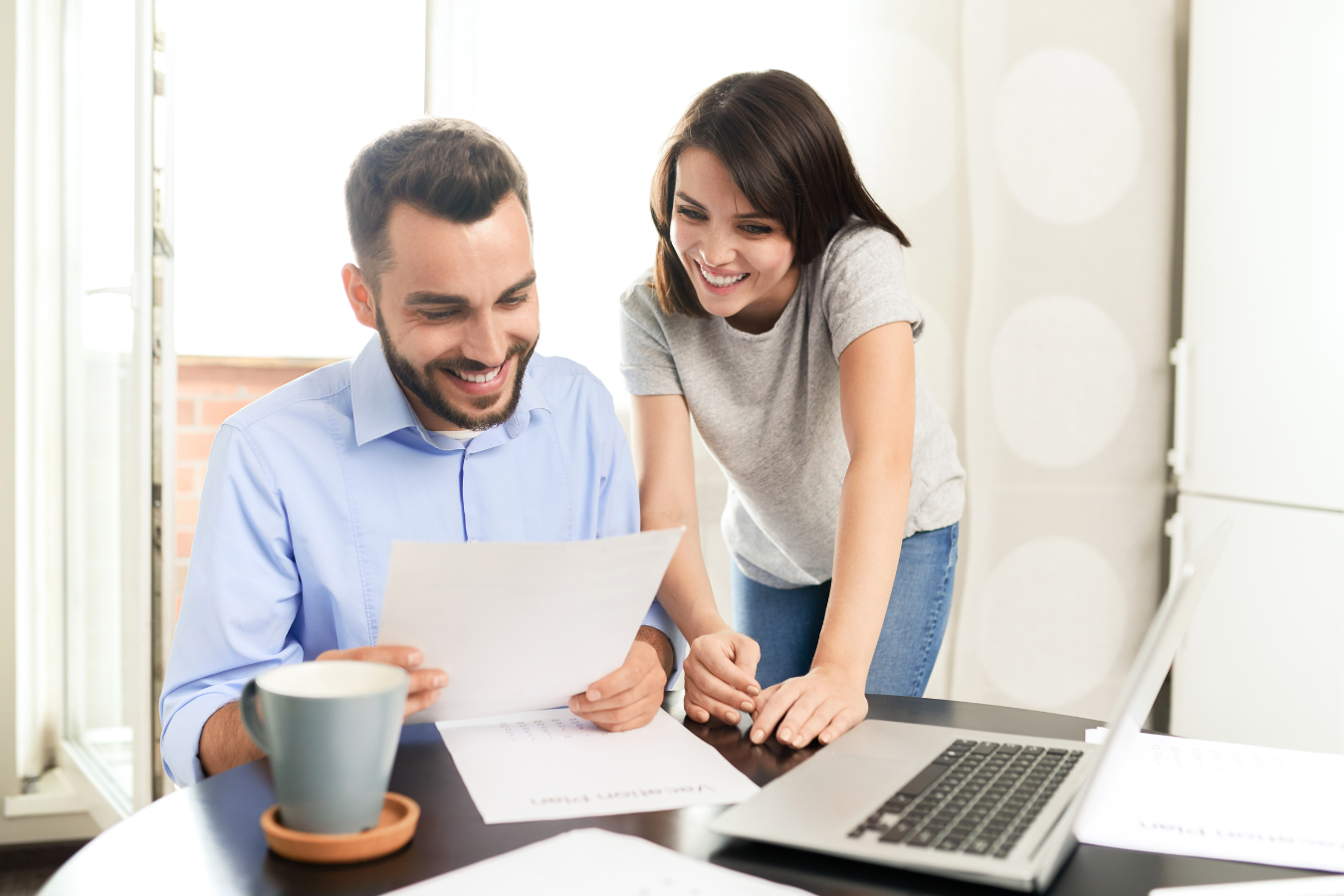 Man and woman looking at papers, smiling, laptop on desk. Natural light.