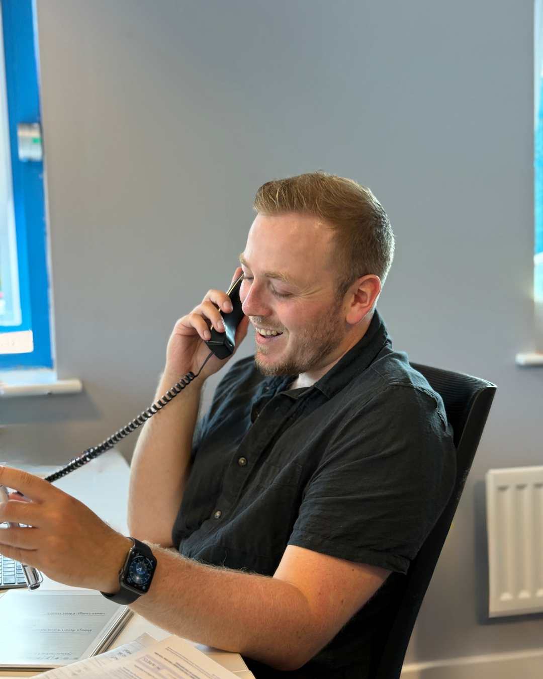 Man on a phone call, smiling, in an office setting. Wearing a black shirt, watch, near a window.