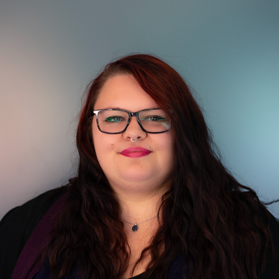 Woman with red hair, glasses, and a nose ring smiles; against a gradient background.