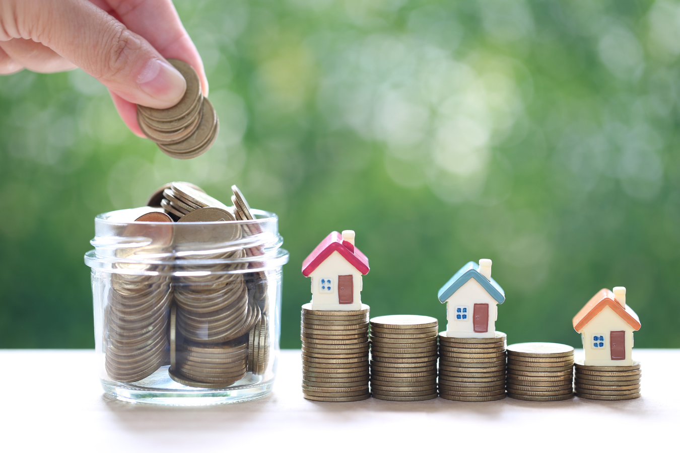 Hand adding coins to a jar, with miniature houses atop stacks of coins.