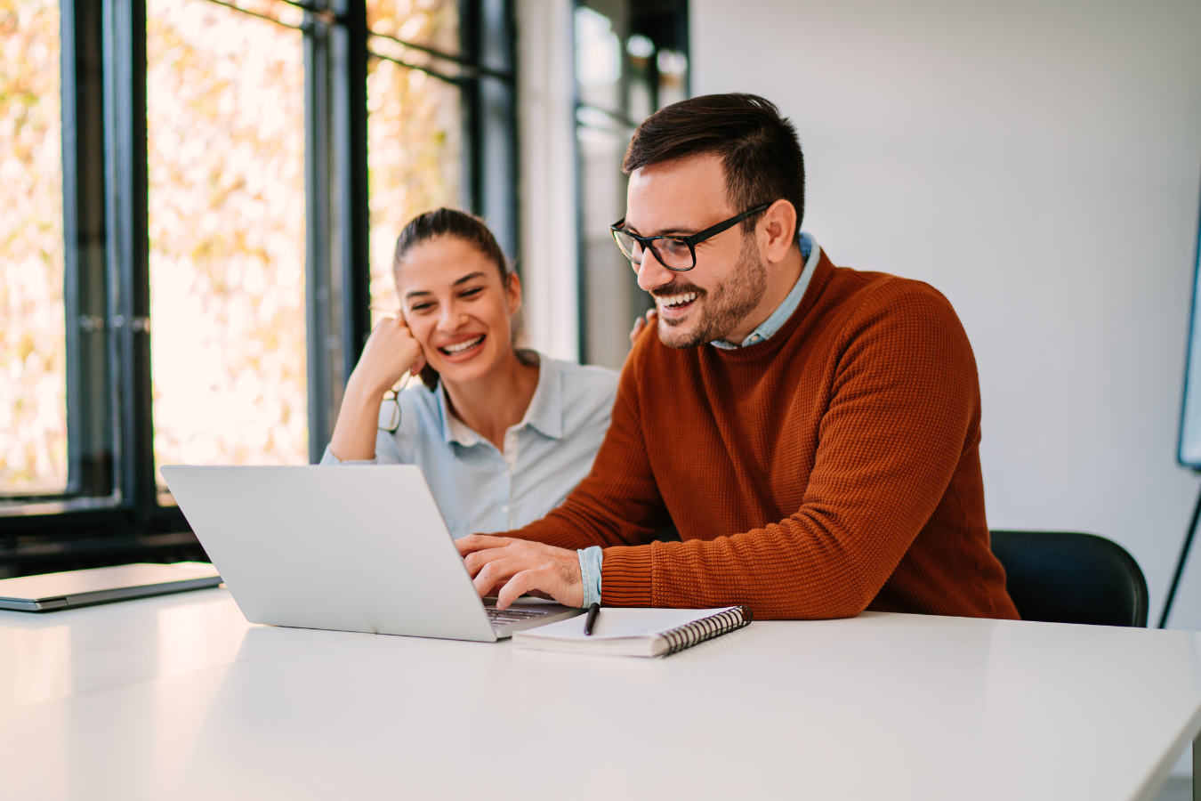 Man and woman smiling at a laptop. The man types, wearing glasses and a brown sweater. Bright, modern office setting.