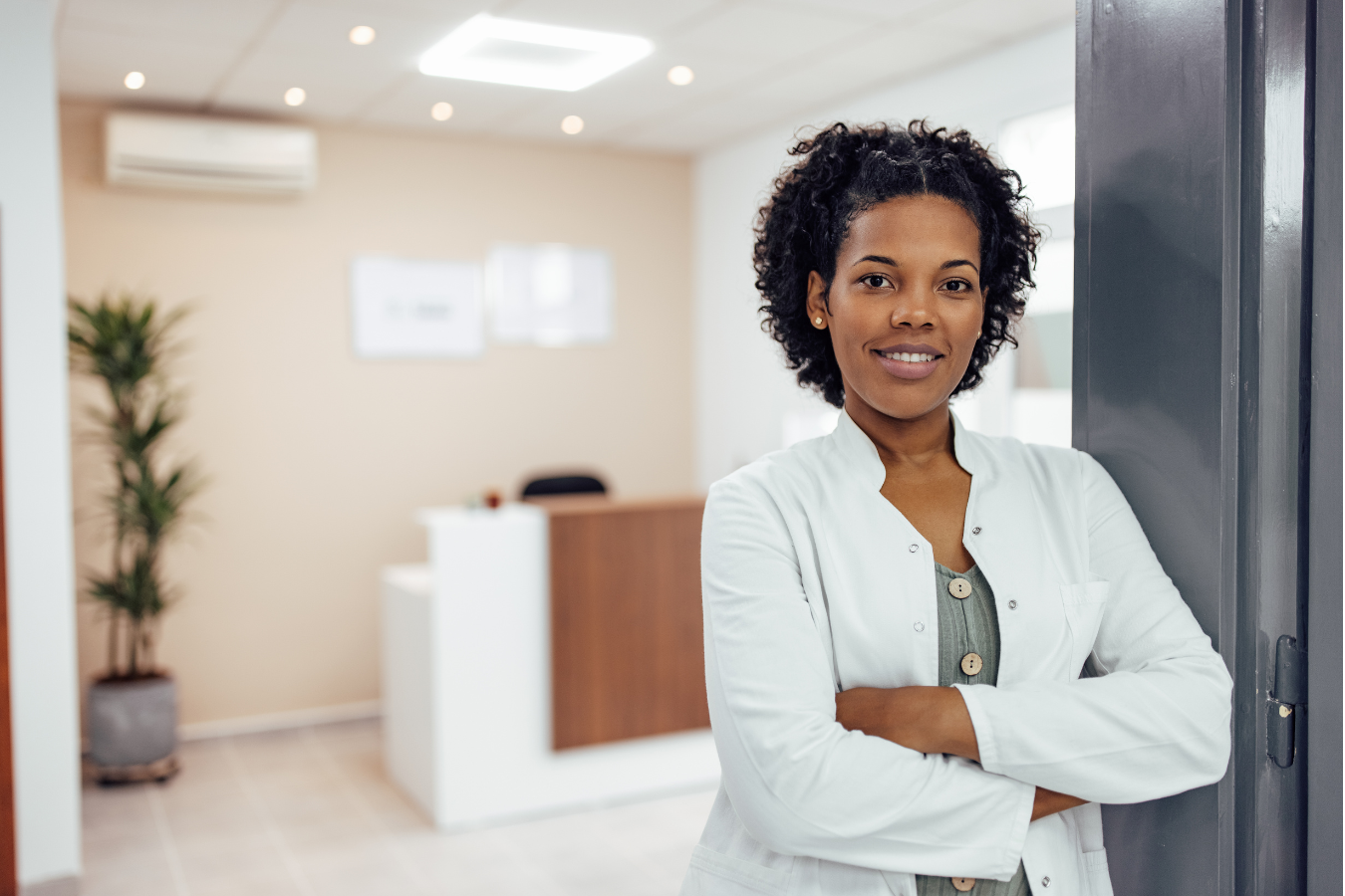 Woman with arms crossed, smiling in an office setting. She wears a white coat, standing by an open door.