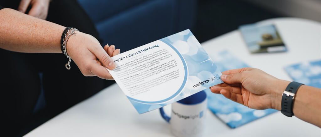Person handing a blue brochure to another person. White table and coffee mug.