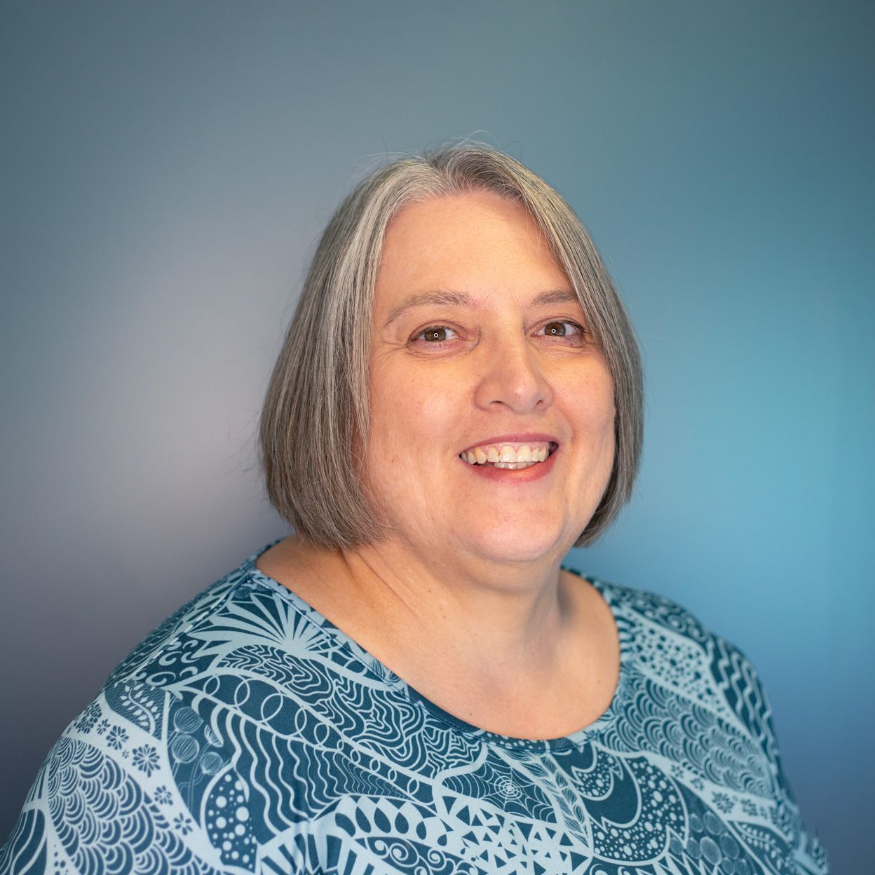 Woman with short gray hair smiles, wearing a blue patterned shirt against a blue gradient background.