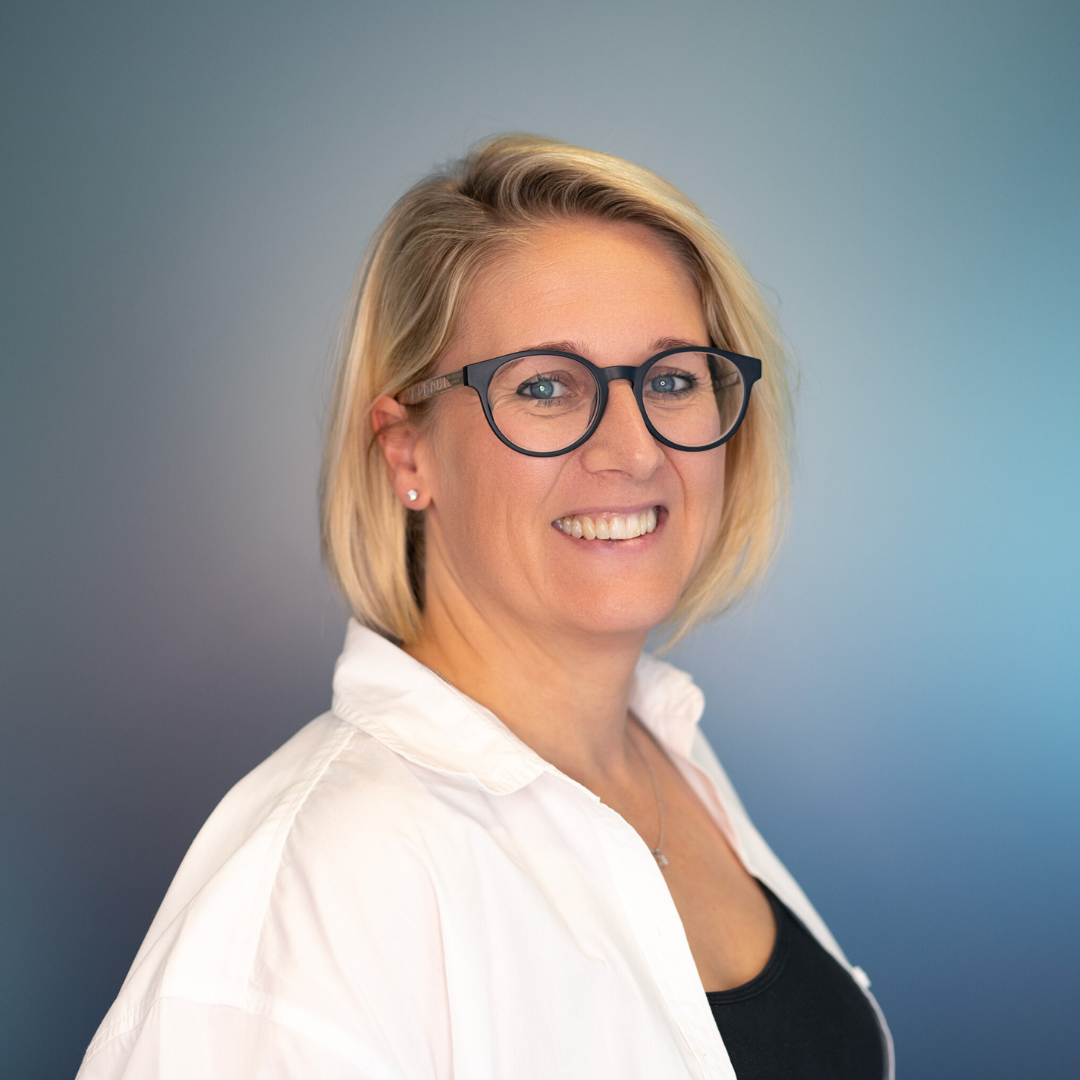 Blonde woman with glasses smiles, wearing a white shirt over a black top, in a studio with blue background.