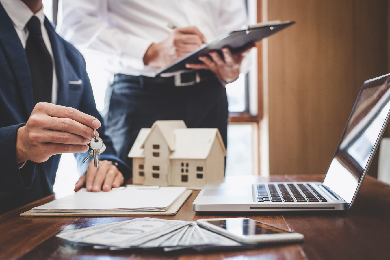 Real estate agent hands keys to a person, small house model, laptop, paperwork, and cash on table.