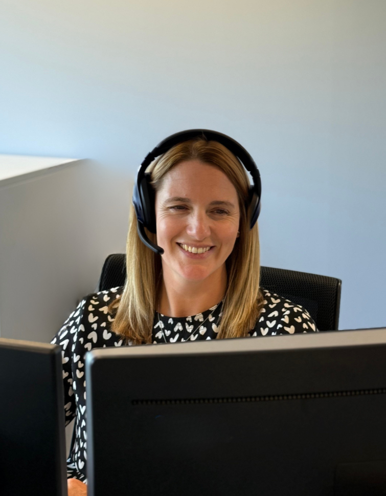 Woman wearing a headset smiles while sitting at a desk in front of a computer.