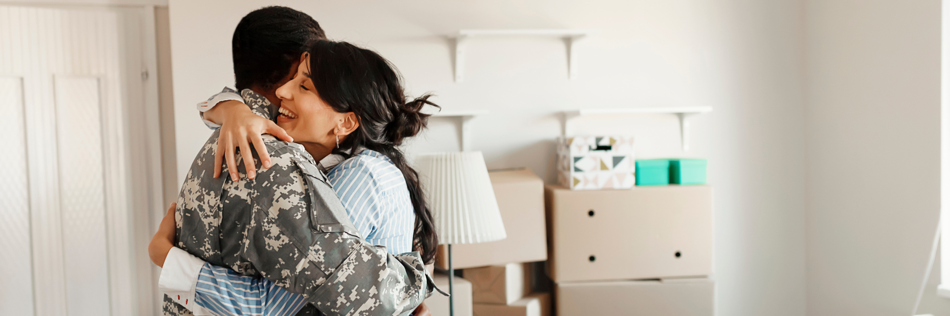 A soldier in camouflage embraces a woman in a new home. Moving boxes are in the background.