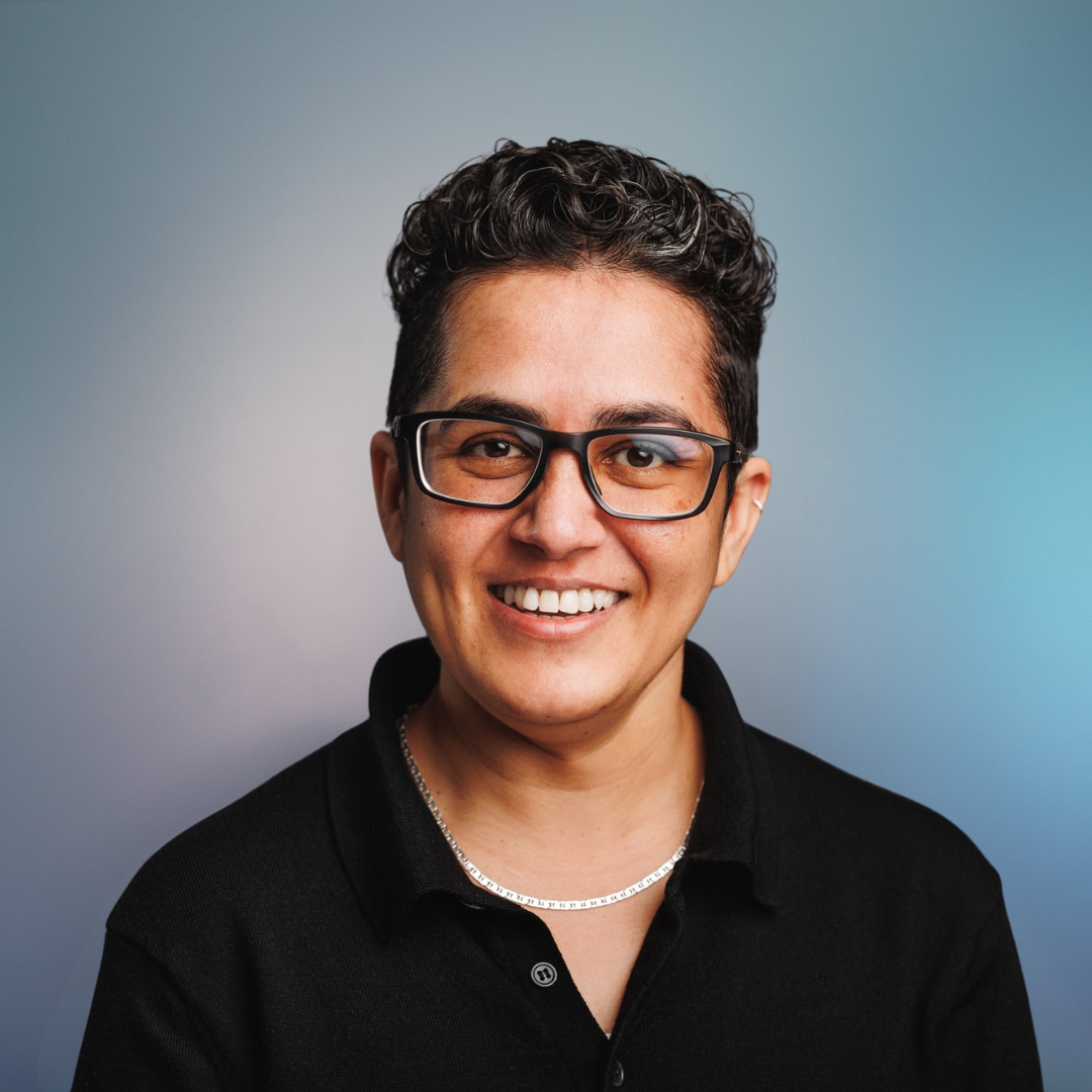 Smiling person with glasses, dark hair, and black shirt, posing against a blurred blue and gray background.