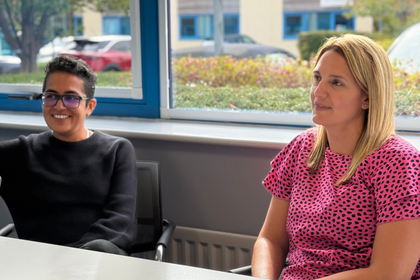 Woman in black blazer laughs, seated on blue couch with two others. Meeting indoors.