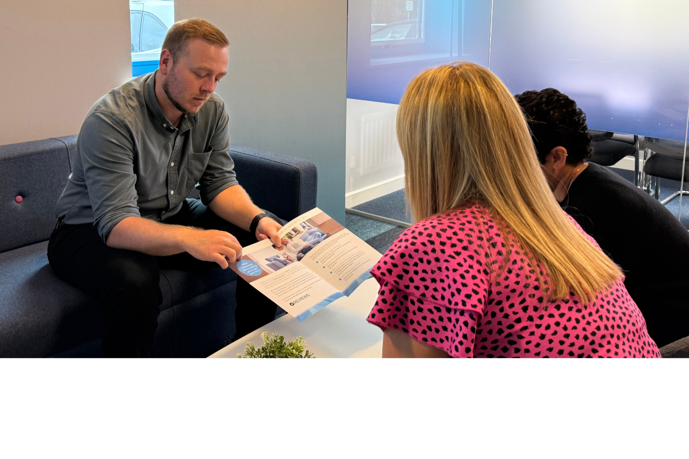 A woman in a blazer smiles, speaking to two people seated on a blue couch.