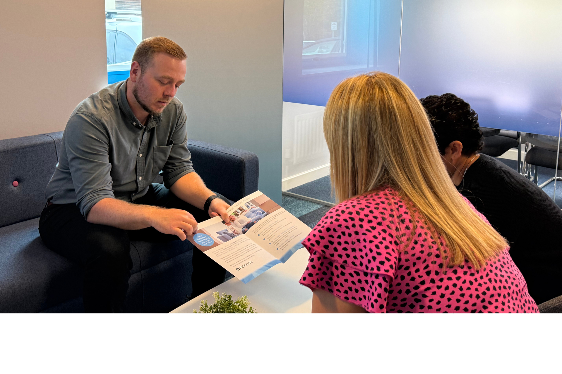 Woman in black blazer laughs, seated on blue couch with two others. Meeting indoors.