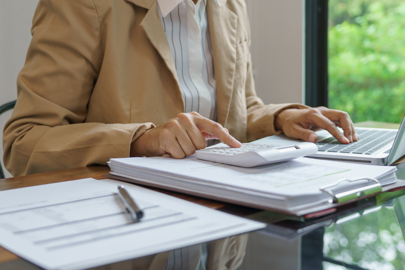 Person in a tan blazer calculates with a calculator, working with documents, a laptop, pen, and clipboard on a desk.