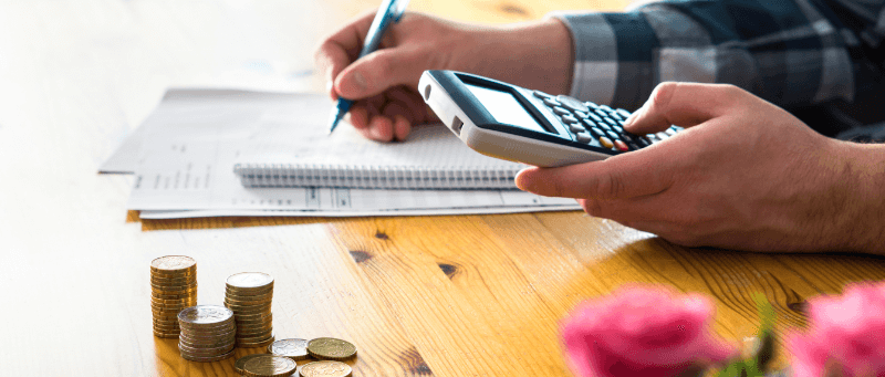 Person calculating finances with a calculator and pen. Stacks of coins and pink flowers on a wooden table.