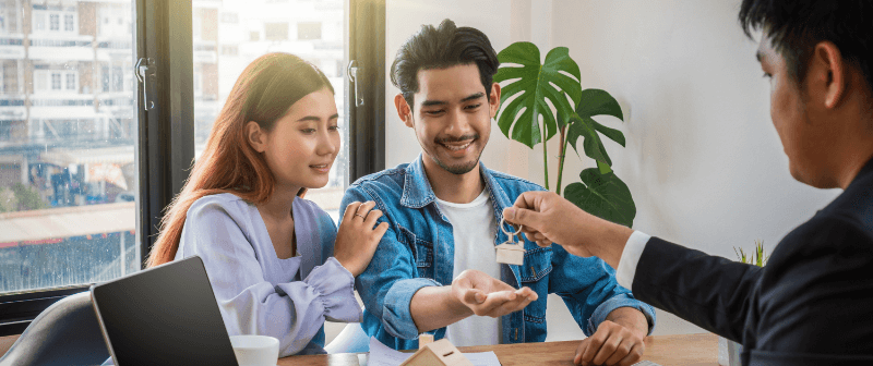 Real estate agent handing keys to a smiling couple at a table.
