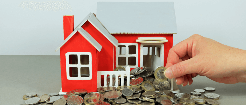 A hand placing a coin near a model of a red house surrounded by coins.