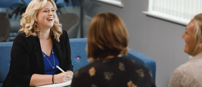 Woman in black blazer laughs, seated on blue couch with two others. Meeting indoors.