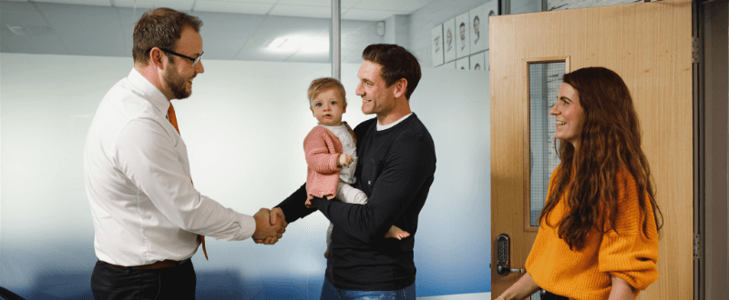 Man shaking hands with another man, holding a baby. Woman standing by a door.