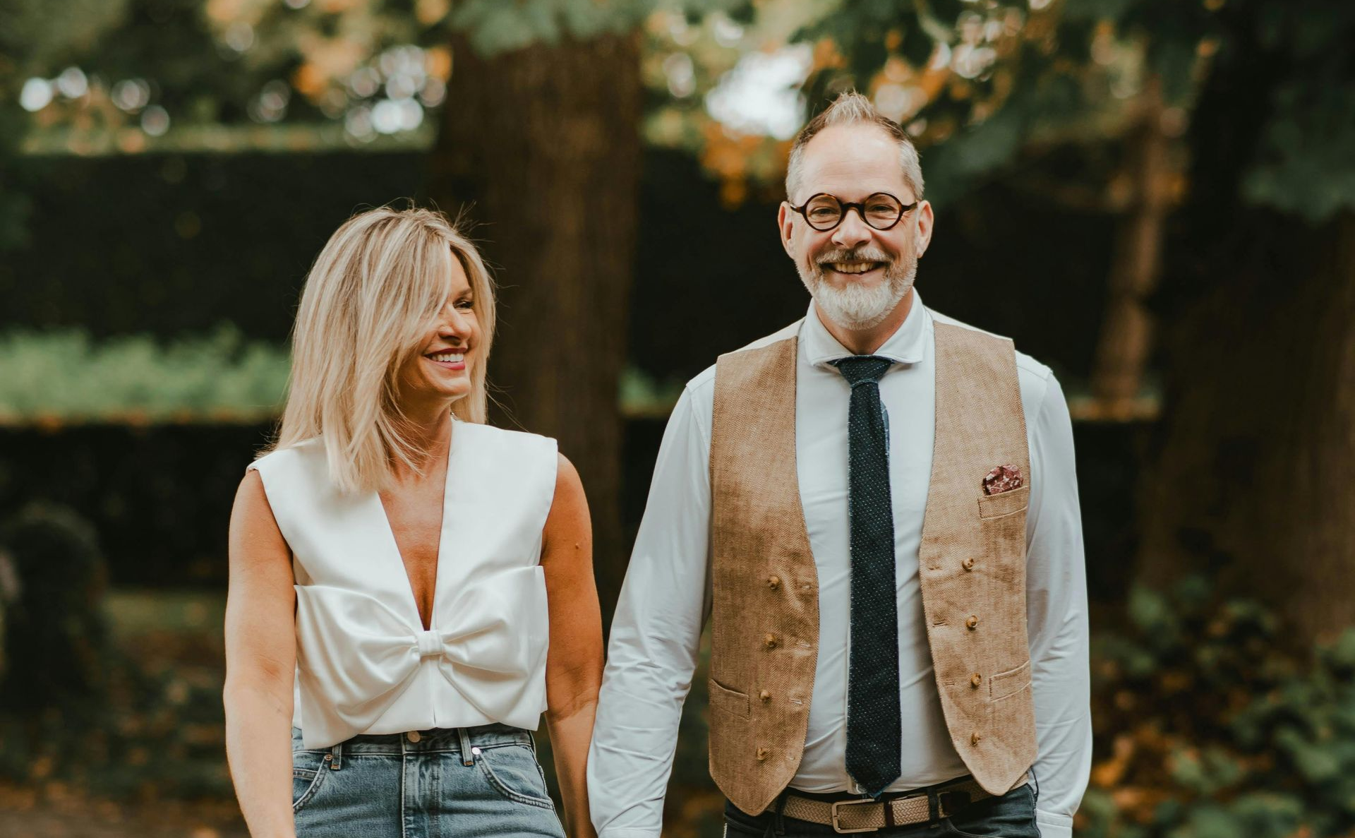 Couple walking, holding hands, smiling; woman in white top and jeans, man in vest and tie, outdoors.