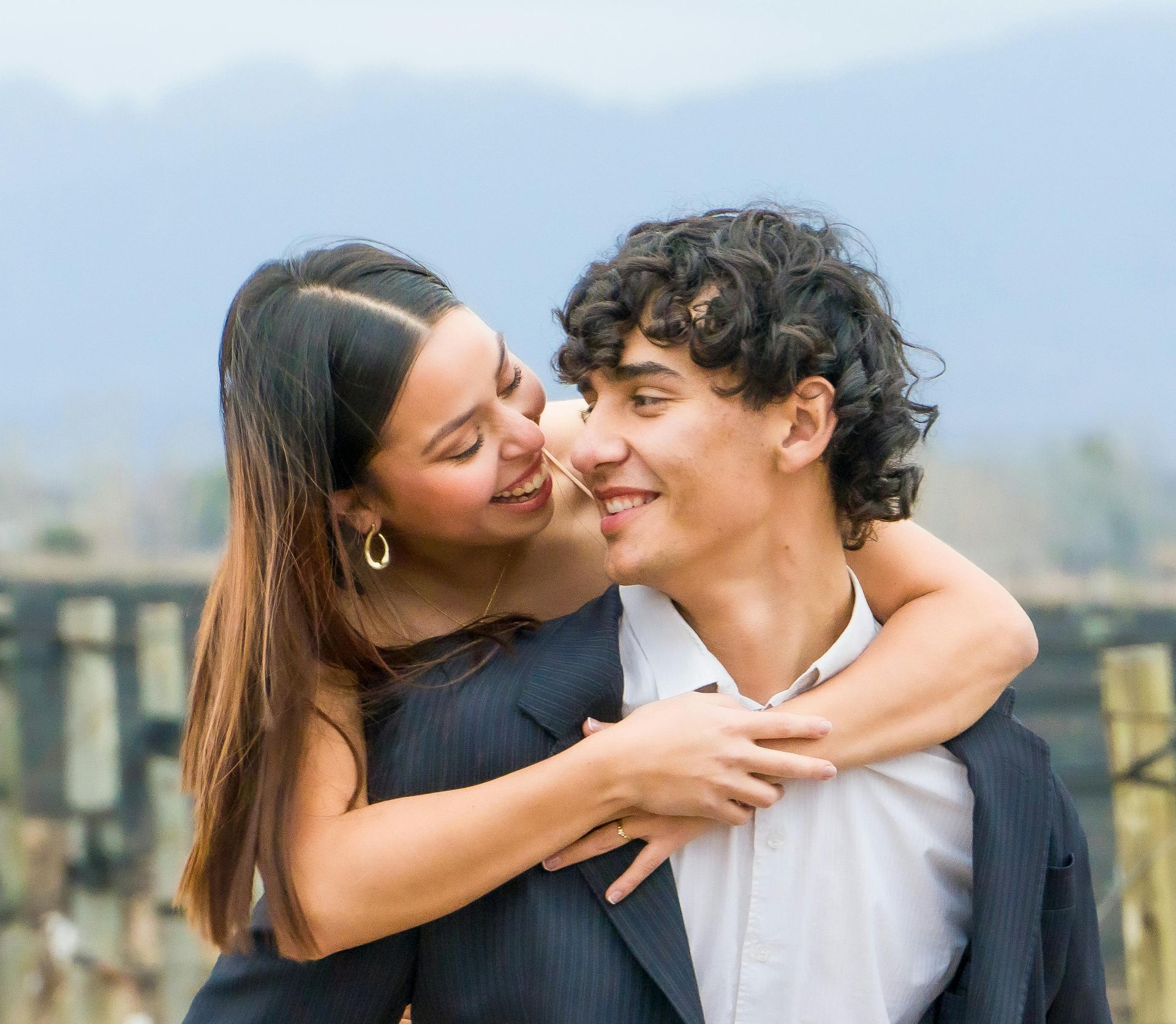 Woman hugs man from behind, both smiling. They are outdoors, with a blurred mountain backdrop.