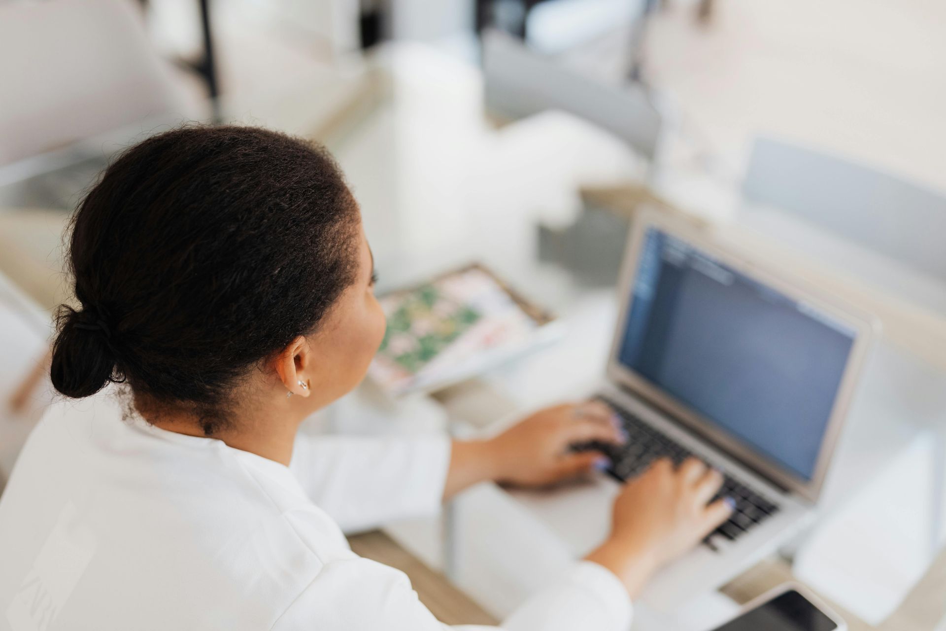 Woman with dark hair types on a laptop at a table; blurred background.