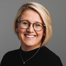 Woman with blonde bob, glasses, smiling. Wearing black sweater, silver necklace. Gray background.