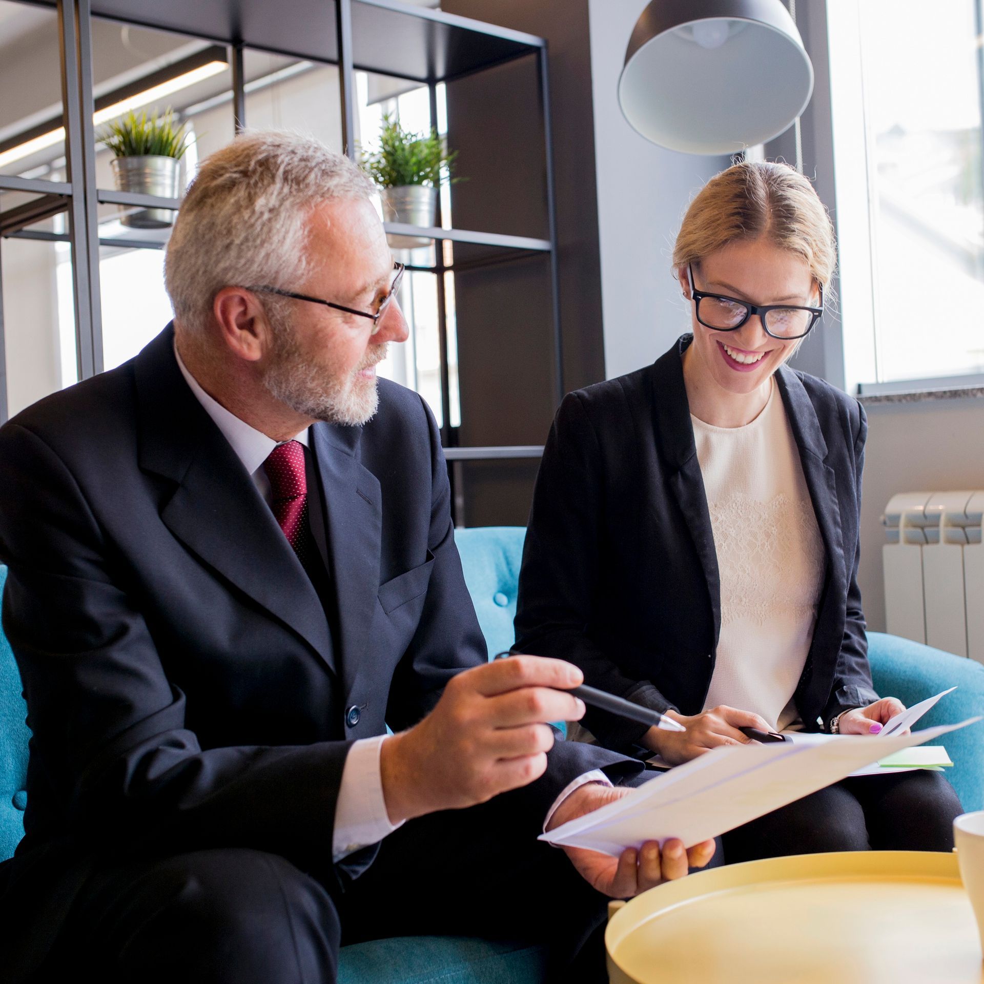 Two people in business attire reviewing documents on a blue couch, discussing.